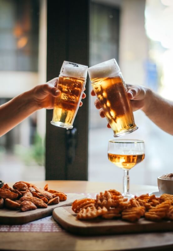 Two people clinking glasses of beer over appetizers and drinks at a restaurant